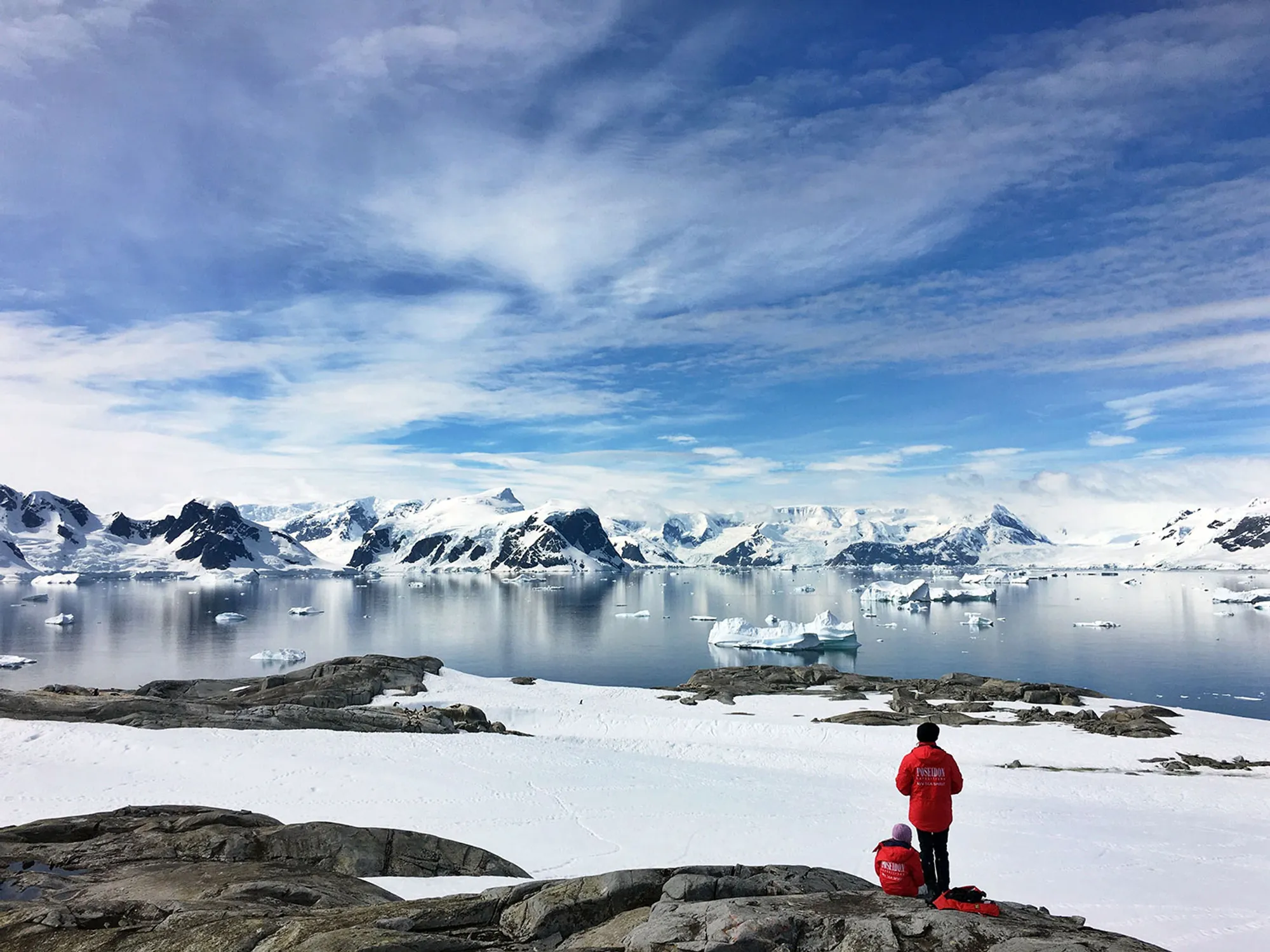 A sweeping photograph of Antarctica's coastline — sheer ice cliffs meeting a calm, mirror-like sea under low polar light.