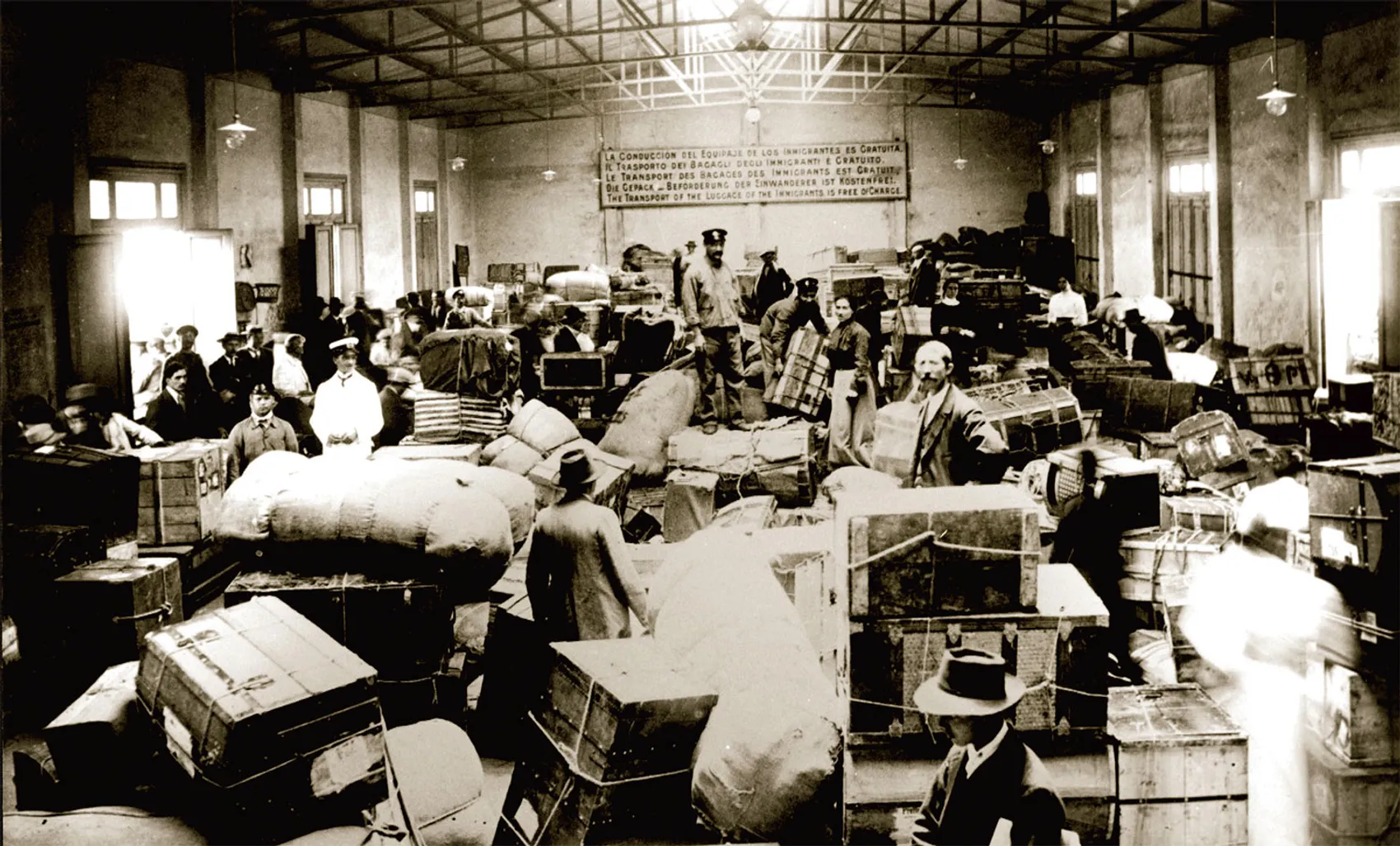 The interior of a vast baggage hall packed with immigrant trunks, sacks, and wooden chests in neatly stacked rows.