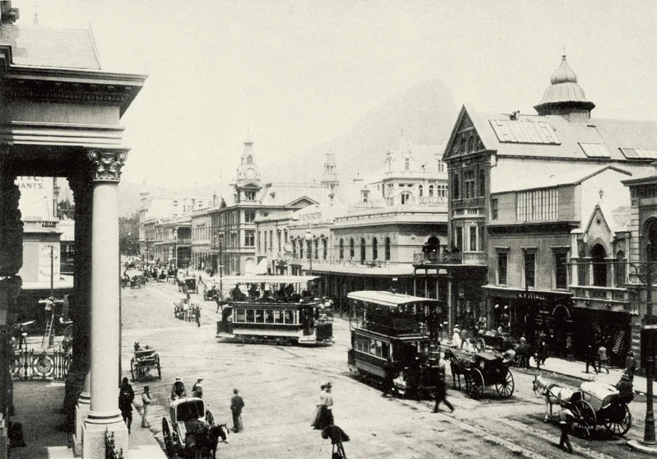 A black-and-white photograph of a busy Cape Town street in the early 1900s, with horse carts, shopfronts, and Table Mountain in the background.