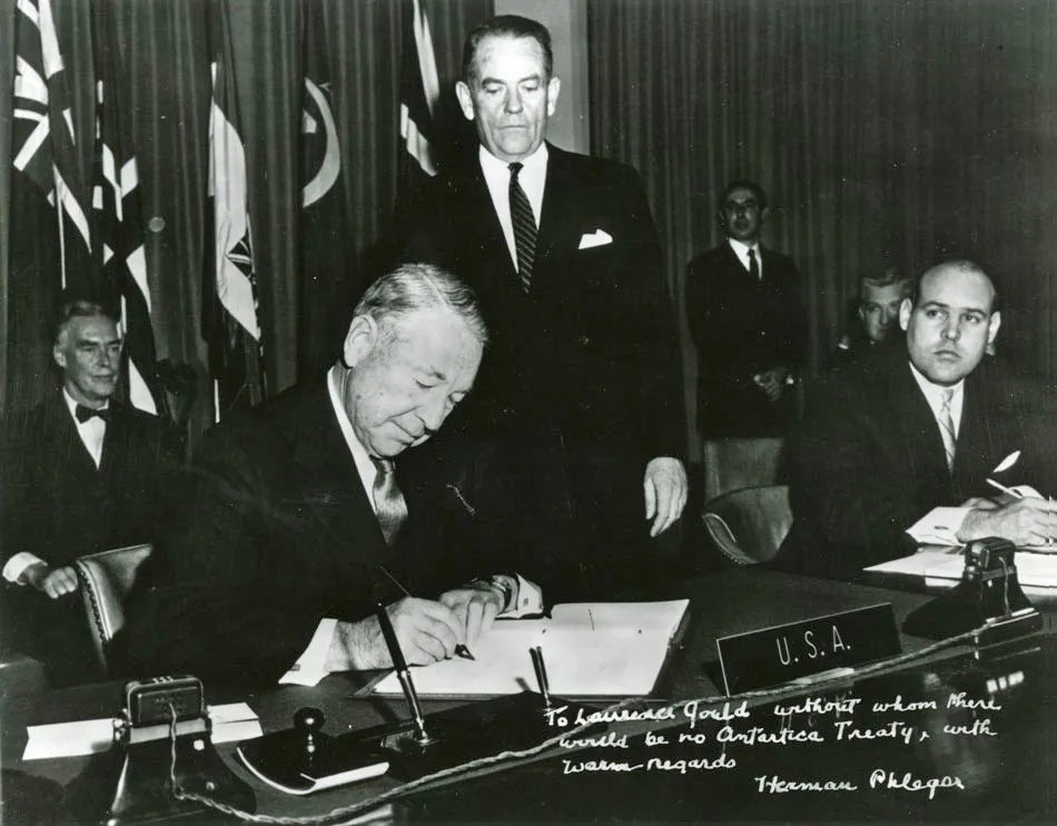 A black-and-white photograph of Herman Phleger seated at a long table, pen in hand, signing the Antarctic Treaty on behalf of the United States in 1959, with officials standing behind him.
