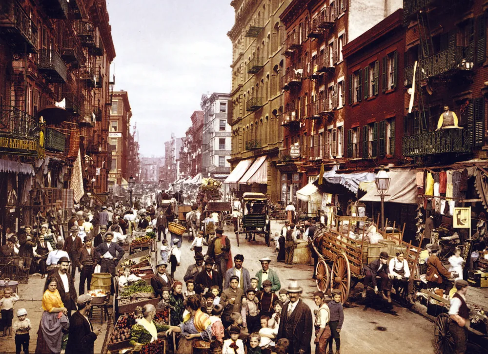 A colour-tinted photograph of New York's Mulberry Street in 1900, packed with pushcarts, shoppers, and the tenement buildings of the Lower East Side.