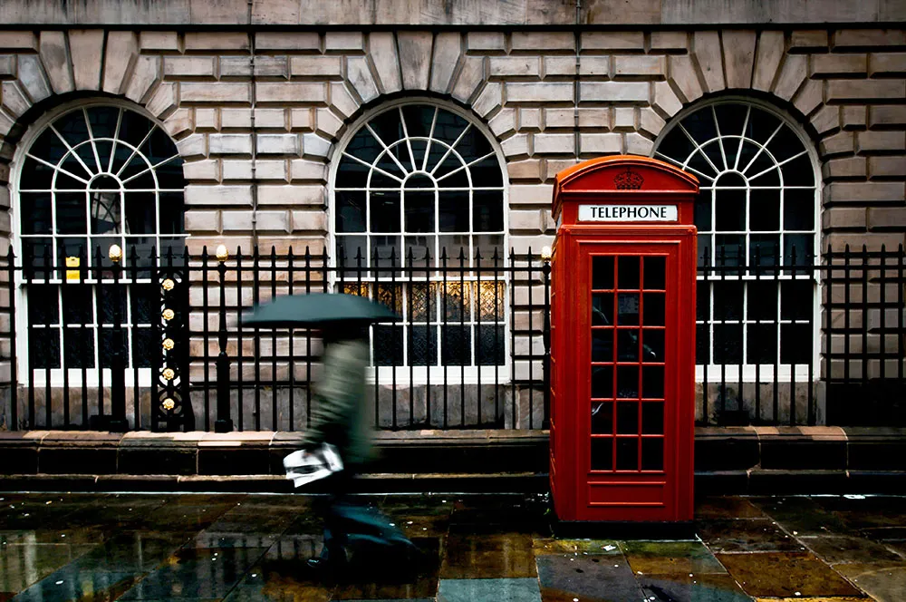 A single iconic red telephone box standing on a London street corner, with traditional buildings behind it.